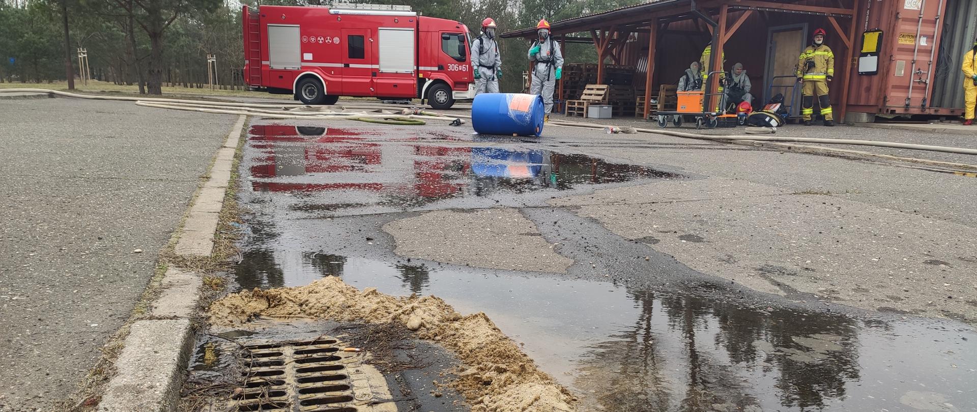dwóch strażaków w szarych ubraniach i w maskach z powietrzem stoi przed niebieską leżącą beczką, za nimi czerwony samochód strażacki i wiata drewniana pod którą stoją strażacy