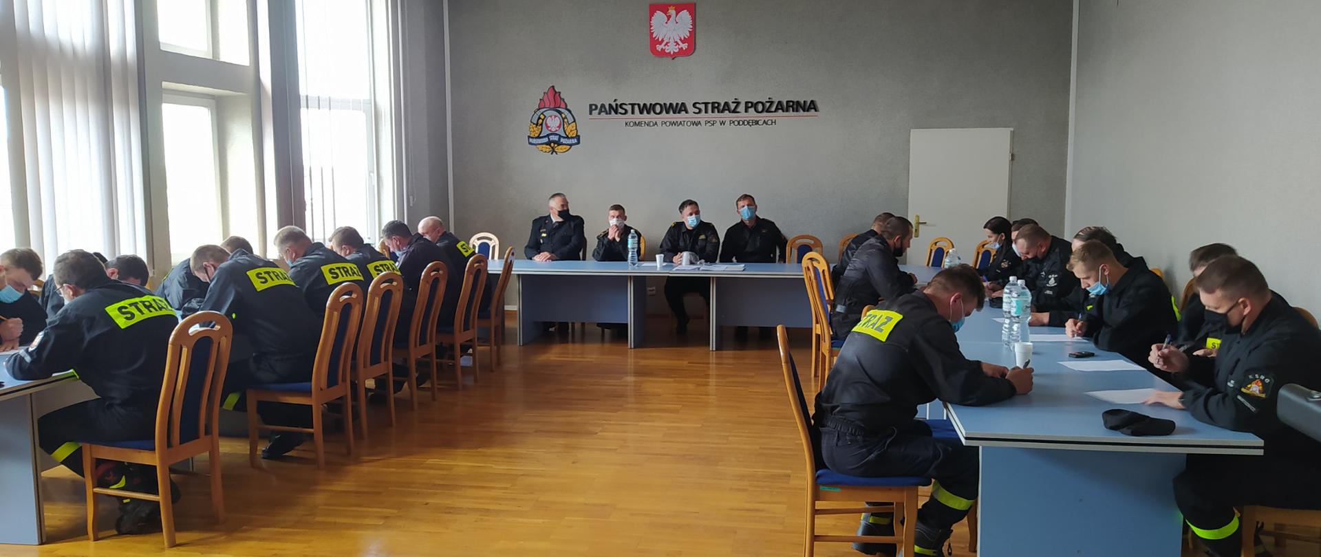 Na sali członkowie OSP piszą egzamin. Są pochyleni nad kartkami. Na szczycie sali siedzą czterej członkowie komisji egzaminacyjnej . Sala ma szare ściany a po lewej stronie na całej szerokości są okna. Nad Komisją na ścianie wisi godło polski a poniżej logo PSP i napis Państwowa Straż Pożarna Komenda Powiatowa PSP w Poddębicach