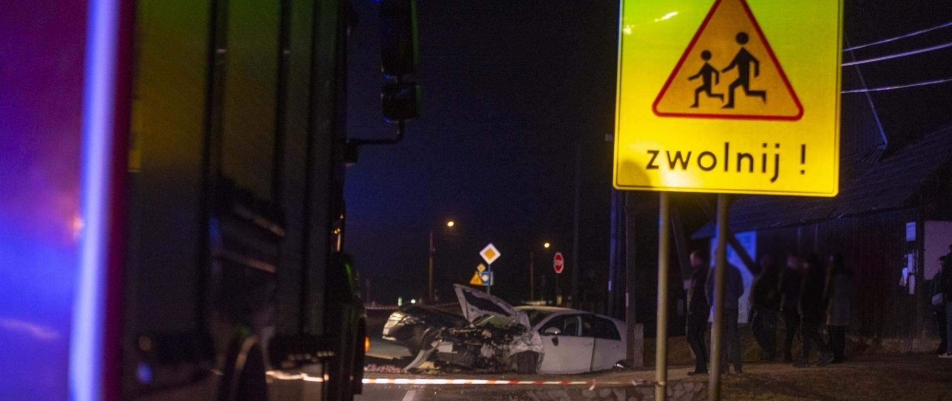 Zdjęcie wykonane w nocy. W tle dwa rozbite samochody znajdujące się w rowie, na pierwszym planie bok samochodu pożarniczego i znak drogowy informacyjny na żółtym tle napis „Kierowco zwolnij”