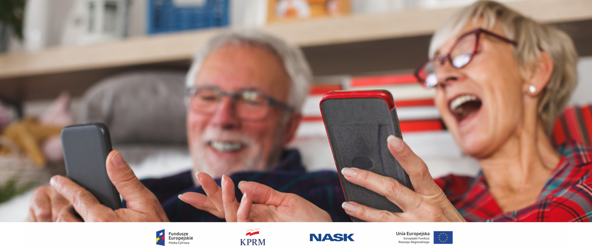 Dwoje seniorów (kobieta i mężczyzna) siedzą na kanapie ze smartfonami w rękach. Oglądają coś zabawnego - oboje się śmieją, wskazując na ekrany urządzeń.