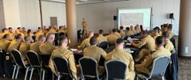 djęcie przedstawia dużą grupę strażaków w piaskowych mundurach służbowych, biorących udział w konferencji lub szkoleniu teoretycznym w przestronnej, eleganckiej sali konferencyjnej. Uczestnicy siedzą przy stołach ustawionych w rzędach, zwróceni tyłem do obiektywu, i uważnie obserwują prezentację wyświetlaną na dużym ekranie projekcyjnym w głębi sali. Prelegent, również ubrany w piaskowy mundur, stoi obok ekranu i omawia schemat organizacyjny widoczny na slajdzie. Wnętrze sali charakteryzuje się nowoczesnym designem, z podwieszanym sufitem, licznymi punktami świetlnymi oraz masywnymi kolumnami wsporczymi. Na stołach widoczne są notatniki, dokumenty oraz butelki z wodą, co podkreśla profesjonalny i oficjalny charakter spotkania kadry dowódczej Państwowej Straży Pożarnej. Zdjęcie dokumentuje proces doskonalenia zawodowego i wymiany doświadczeń w strukturach straży.