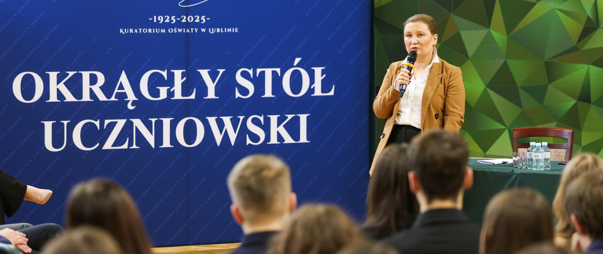 Wiceminister Paulina Piechna-Więckiewicz, ubrana w brązową marynarkę, stoi przed publicznością, przemawiając do mikrofonu. Za nią widoczne są dwa banery: niebieski z logo i napisem "OKRĄGŁY STÓŁ UCZNIOWSKI" oraz zielono-czarny z nazwą szkoły "LICEUM „ZAPORY”"