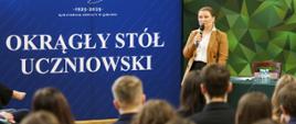Wiceminister Paulina Piechna-Więckiewicz, ubrana w brązową marynarkę, stoi przed publicznością, przemawiając do mikrofonu. Za nią widoczne są dwa banery: niebieski z logo i napisem "OKRĄGŁY STÓŁ UCZNIOWSKI" oraz zielono-czarny z nazwą szkoły "LICEUM „ZAPORY”"
