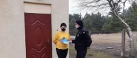 Zdjęcie przedstawia strażaka OSP, który wręcza kobiecie ulotkę dotyczącą szczepień przeciwko COVID-19. Zdjęcie wykonane jest przed wejściem do budynku mieszkalnego. W tle zdjęcia po prawej stronie widać rosnący las za ogrodzeniem. 