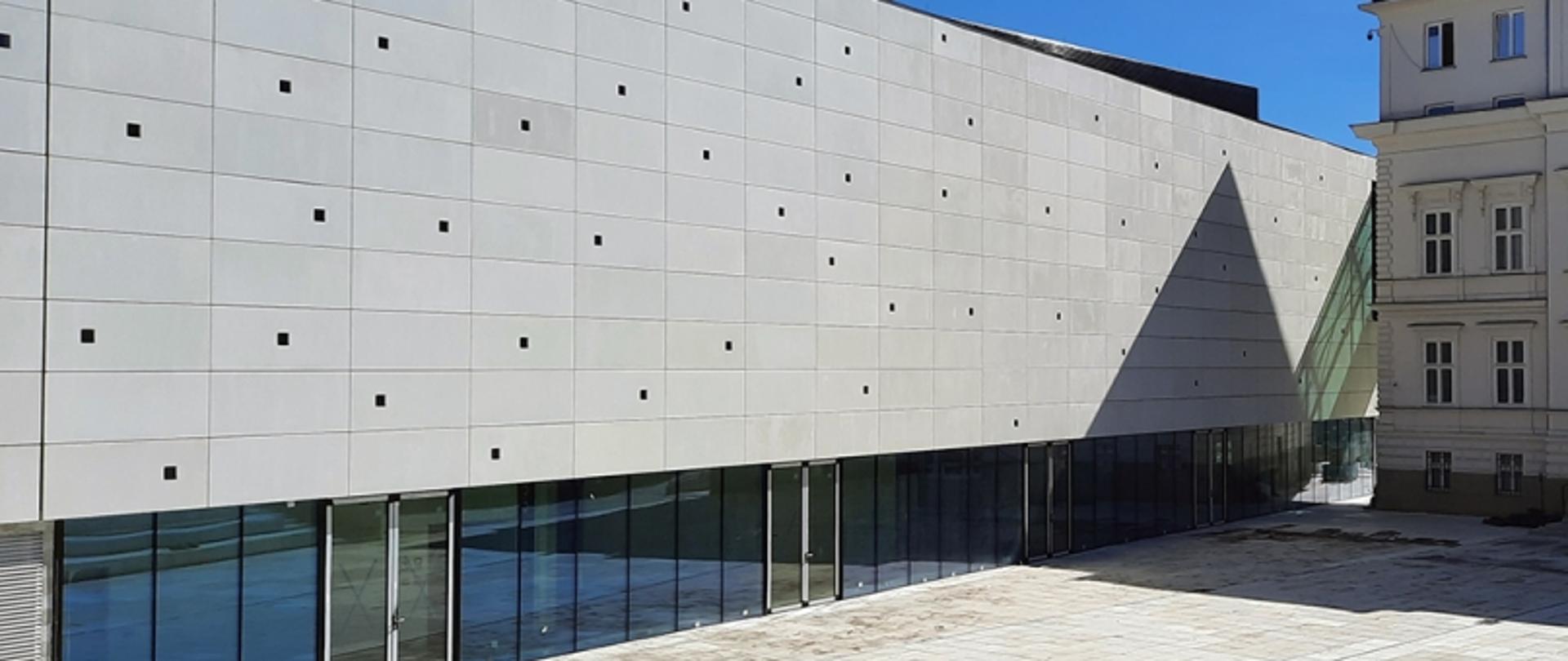 Nowoczesny budynek z siatką małych okien i przeszklonym parterem, obok starszy budynek z wieloma oknami; na pierwszym planie dziedziniec.