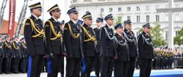 Grupa wyróżnionych funkcjonariuszy PSP oraz druhów OSP stoi w szeregu na placu podczas uroczystości. Na ich mundurach widoczne są świeżo przypięte odznaczenia państwowe.