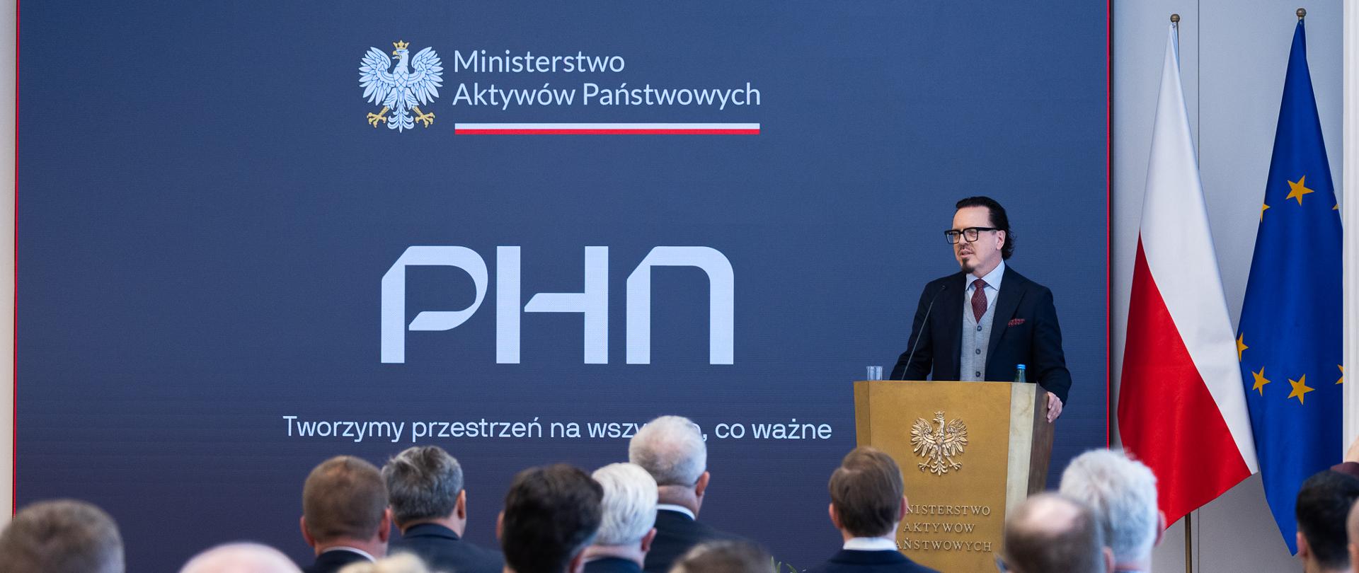 Minister Wojciech Balczun w trakcie wypowiedzi, stoi za mównicą. W tle ścianka z napisem PHN -Tworzymy przestrzeń na wszystko, co ważne. Ujęcie zza publiczności.