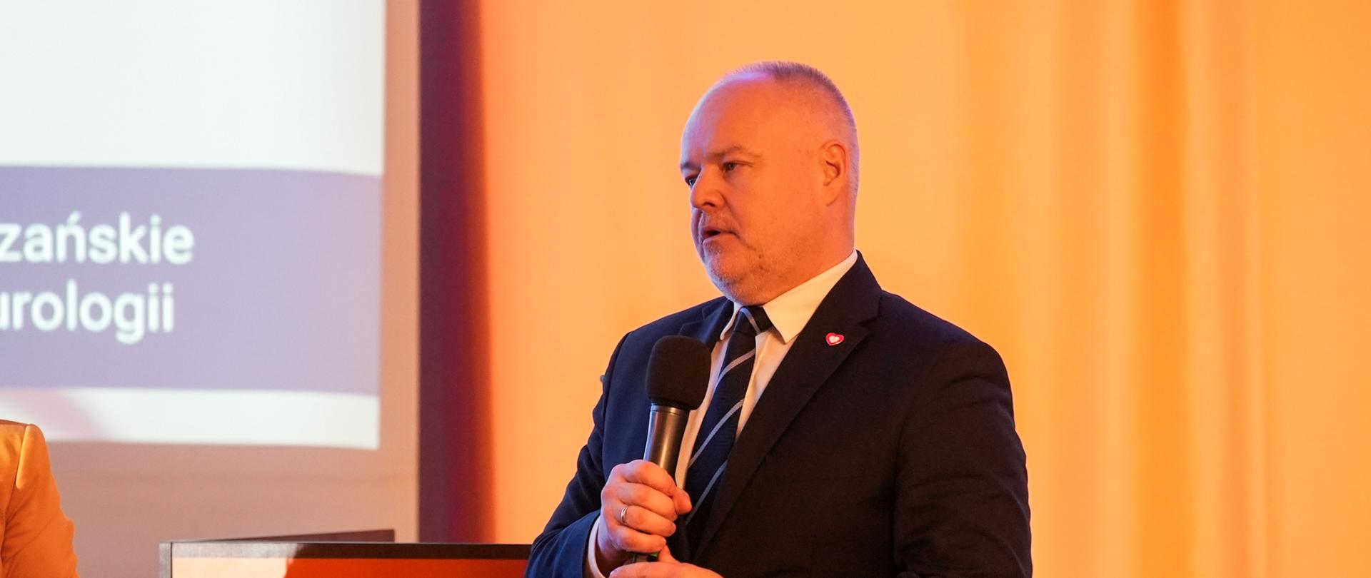 Na sali stoi wiceminister Gzik i mówi do trzymanego w ręku mikrofonu, za nim żółta ściana, z lewej fragment ekranu z napisem XI Zabrzańskie Dni Neurologii.