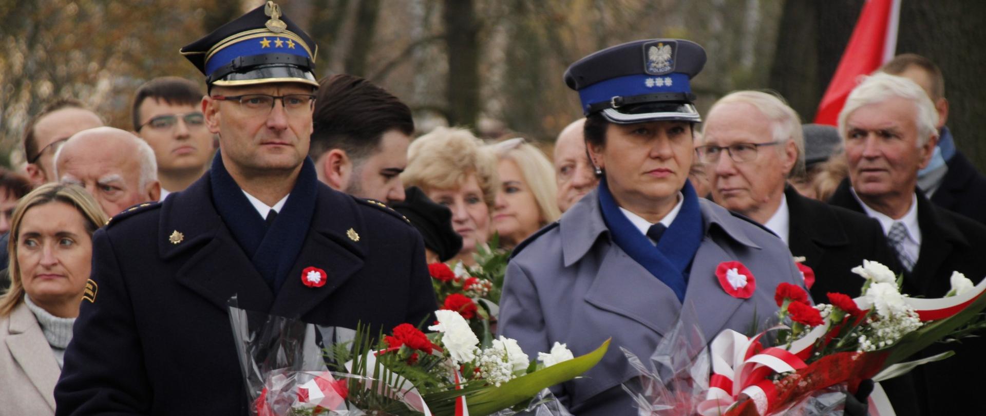 Komendant powiatowy PSP w Żyrardowie st. bryg. Mariusz Tymoszewicz stoi obok Komendanta Powiatowego Policji w Żyrardowie. W rękach trzymają wiązanki kwiatów w kolorze biało- czerwonym