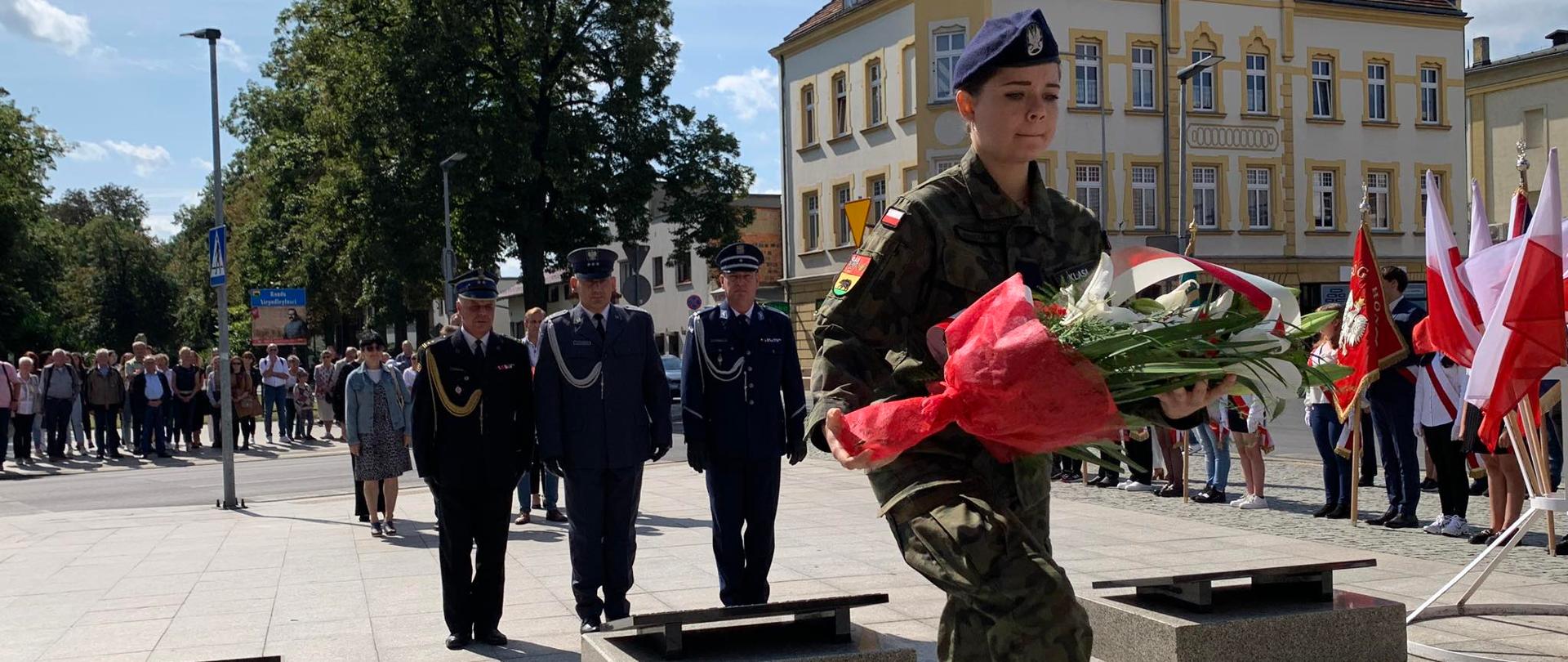 Rocznica wybuchu II WŚ. Zdjęcie przedstawia plac przed pomnikiem Żołnierza Polskiego. Na pierwszym planie uczennica klasy mundurowej niosąca pod pomnik wiązankę kwiatów. Przed tablica znajduje się delegacja służb mundurowych: Państwowej Straży Pożarnej, Służby Więziennej i Policji. Funkcjonariusze występują w umundurowaniu galowym. Z prawej strony stoją w szeregu poczty sztandarowe, z tyłu natomiast pozostałe delegacje oraz mieszkańcy miasta.