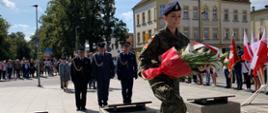 Rocznica wybuchu II WŚ. Zdjęcie przedstawia plac przed pomnikiem Żołnierza Polskiego. Na pierwszym planie uczennica klasy mundurowej niosąca pod pomnik wiązankę kwiatów. Przed tablica znajduje się delegacja służb mundurowych: Państwowej Straży Pożarnej, Służby Więziennej i Policji. Funkcjonariusze występują w umundurowaniu galowym. Z prawej strony stoją w szeregu poczty sztandarowe, z tyłu natomiast pozostałe delegacje oraz mieszkańcy miasta.