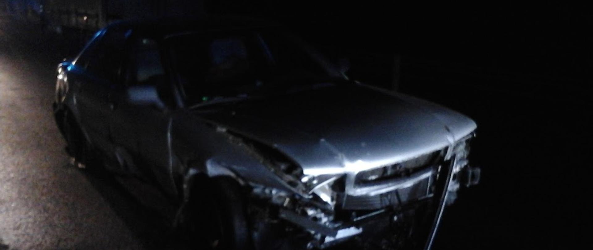 Uszkodzony samochód osobowy stojący na jezdni po kolizji. Uszkodzony przód pojazdu oraz cały bok od strony pasażera.