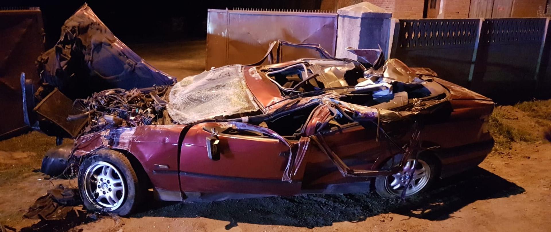 Zgnieciony samochód osobowy w pozycji na kołach, stojący w wjeździe do prywatnej posesji.