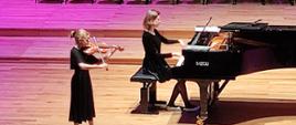 Dziewczynka gra na skrzypcach stojąc po lewej stronie, obok po prawej kobieta gra na fortepianie