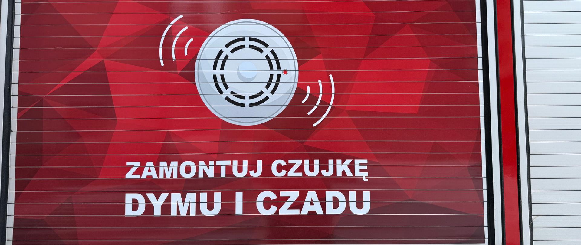 Drzwi schowka wozu strażackiego, pomalowane na czerwono z białymi elementami. Na drzwiach znajduje się duża grafika i tekst w języku polskim.
W górnej części widnieje napis "ZADBAJ O BEZPIECZEŃSTWO". Poniżej znajduje się duży symbol czujnika dymu/czadu, emitującego sygnały, a pod nim tekst "ZAMONTUJ CZUJKĘ DYMU I CZADU". Na samym dole, po prawej stronie, widać mały biały napis "WISS".