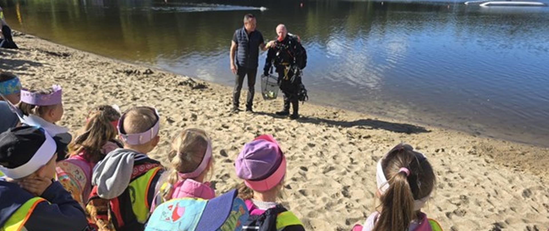 Obraz przedstawia grupę dzieci oraz płetwonurka z osobą przeprowadzającą z nim wywiad. W tle widać jezioro Głębokie.