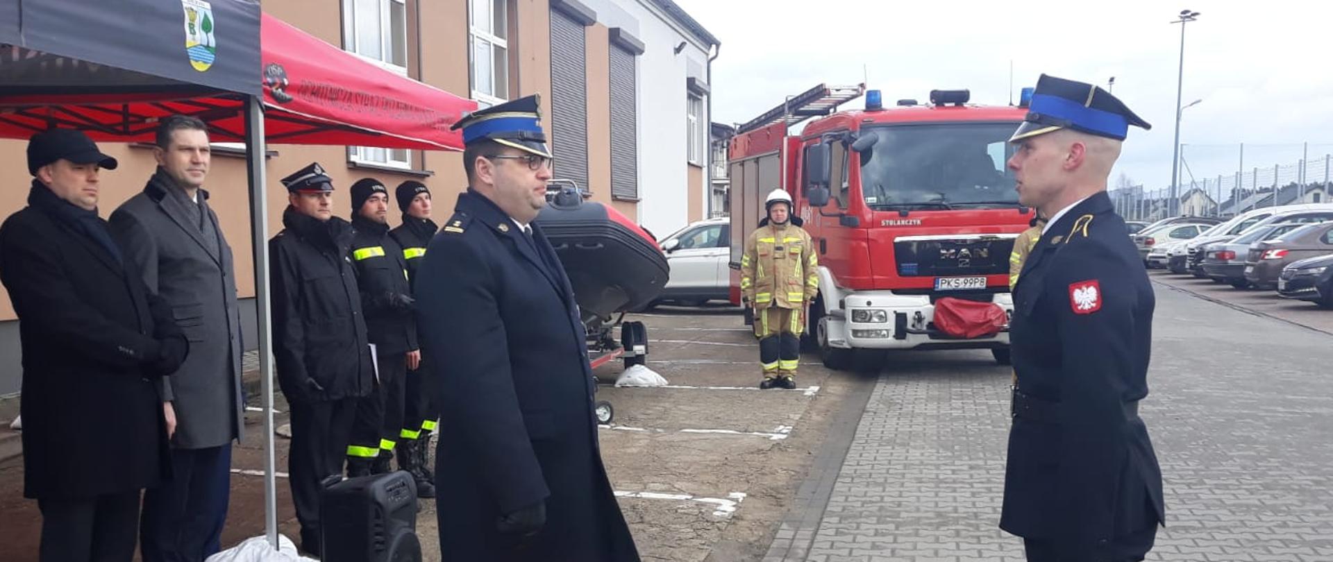 przed remizą strażacką składanie meldunku strażakowi, za nim grupa ludzi pod namiotem, w tle samochód strażacki