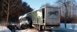 Ciągnik siodłowy IVECO z naczepą po uderzeniu w przydrożny nasyp. Przód pojazdu rozbity. Ciągnik siodłowy obrócony tyłem do kierunku jazdy częściowo wgnieciony w naczepę. Droga oblodzona, pobocze drogi zaśnieżone.