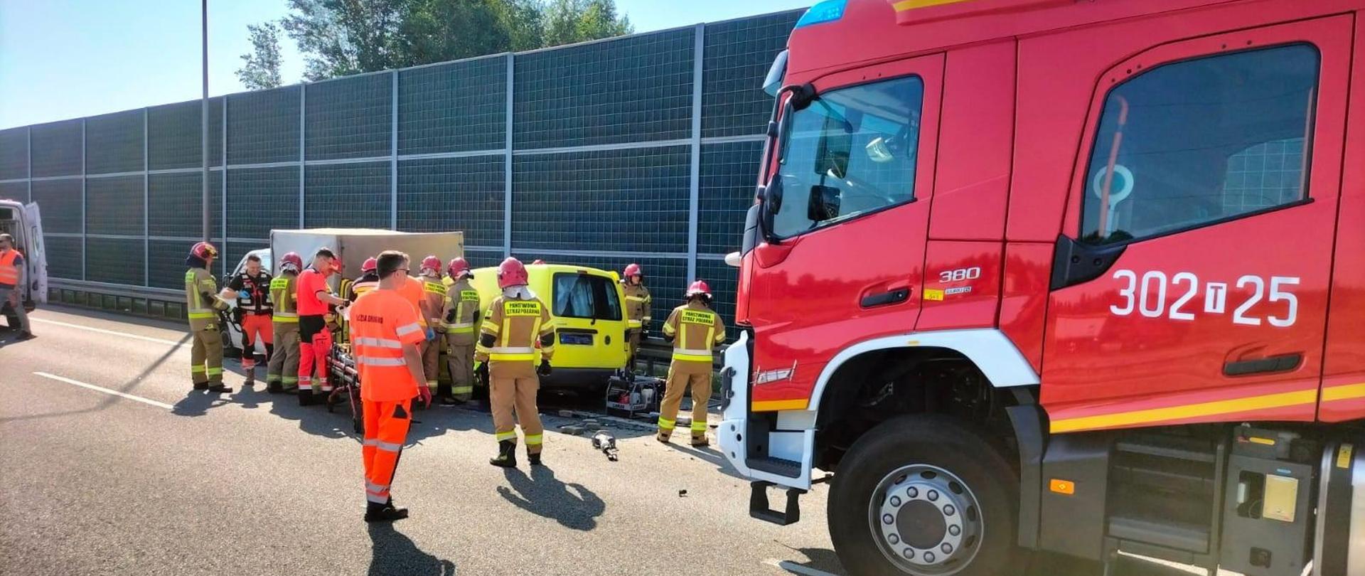 Zdjęcie przedstawia drogę ekspresową S7 i miejsce zderzenia 3 samochodów. Od prawej stoją: samochód pożarniczy i po kolei 3 auta biorące udział w wypadku. Między pojazdami stoją ratownicy medyczni i strażacy.