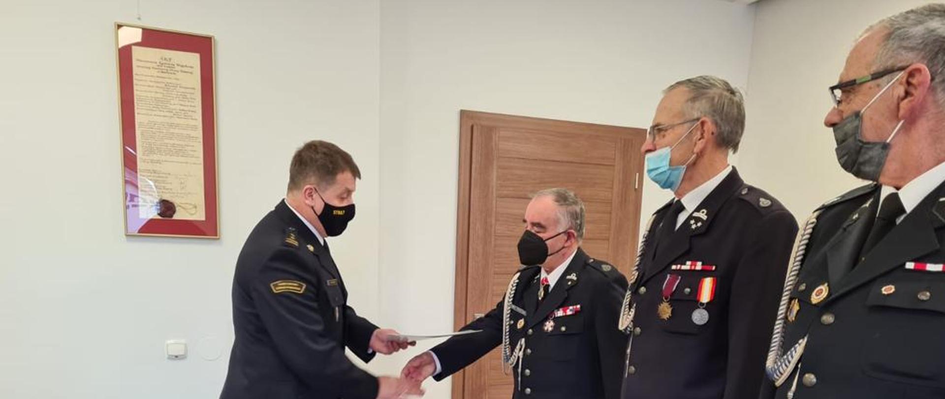 Na zdjęciu na sali konferencyjnej Komendant Powiatowy wręcza Decyzję o przyznaniu świadczenia ratowniczego druhowi OSP na zdjęciu w szeregi trzech strażaków OSP i jeden PSP w granatowych mundurach galowych. 