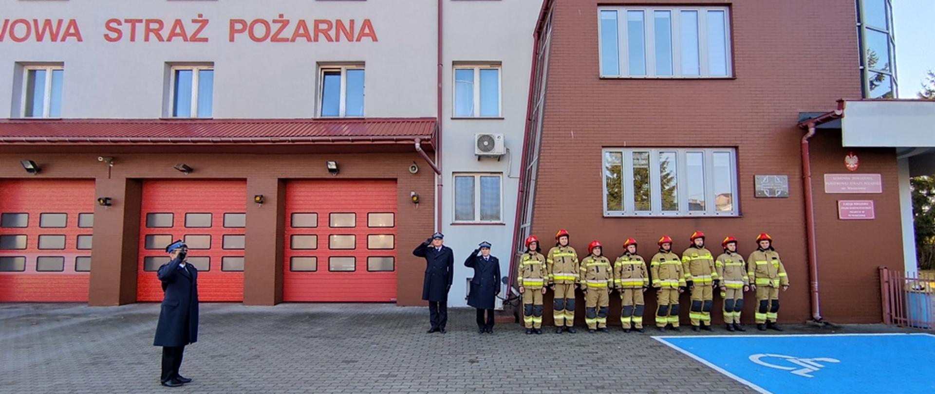Plac przed budynkiem Komendy Powiatowej PSP we Włoszczowie, zmiana służbowa stojąca przed budynkiem w ubraniach specjalnych obok nich kierownictwo włoszczowskiej komendy w umundurowani wyjściowym