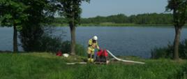Strażak stoi przy motopompie i rozmawia przez radiostację. Od motopompy odchodzi wąż pożarniczy. W tle staw i drzewa.