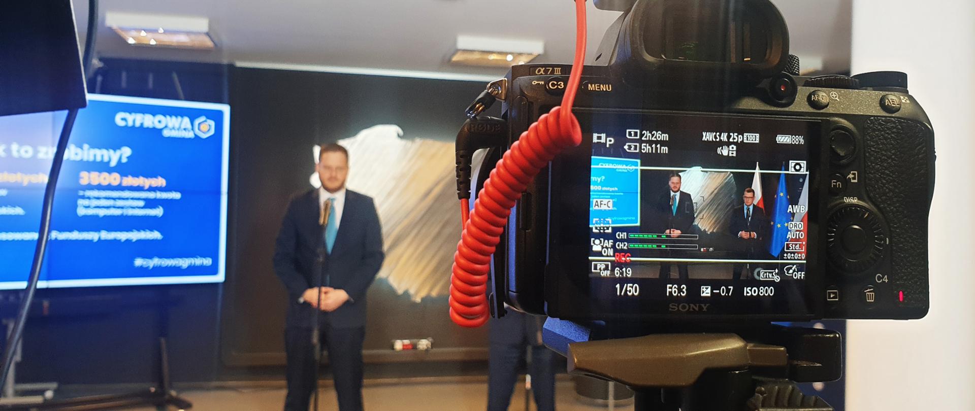 Zdjęcie przedstawiające ministra Janusza Cieszyńskiego podczas konferencji prasowej. Na pierwszym planie widać kadr w obiektywie kamery.