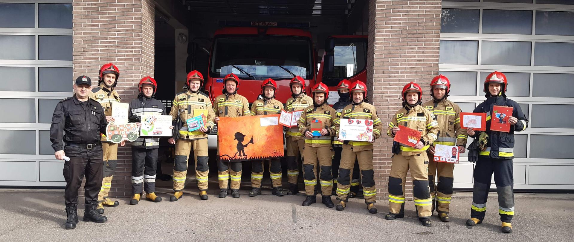 Strażacy w umundurowaniu bojowym trzymają w ręce prace plastyczne otrzymane w prezencie.