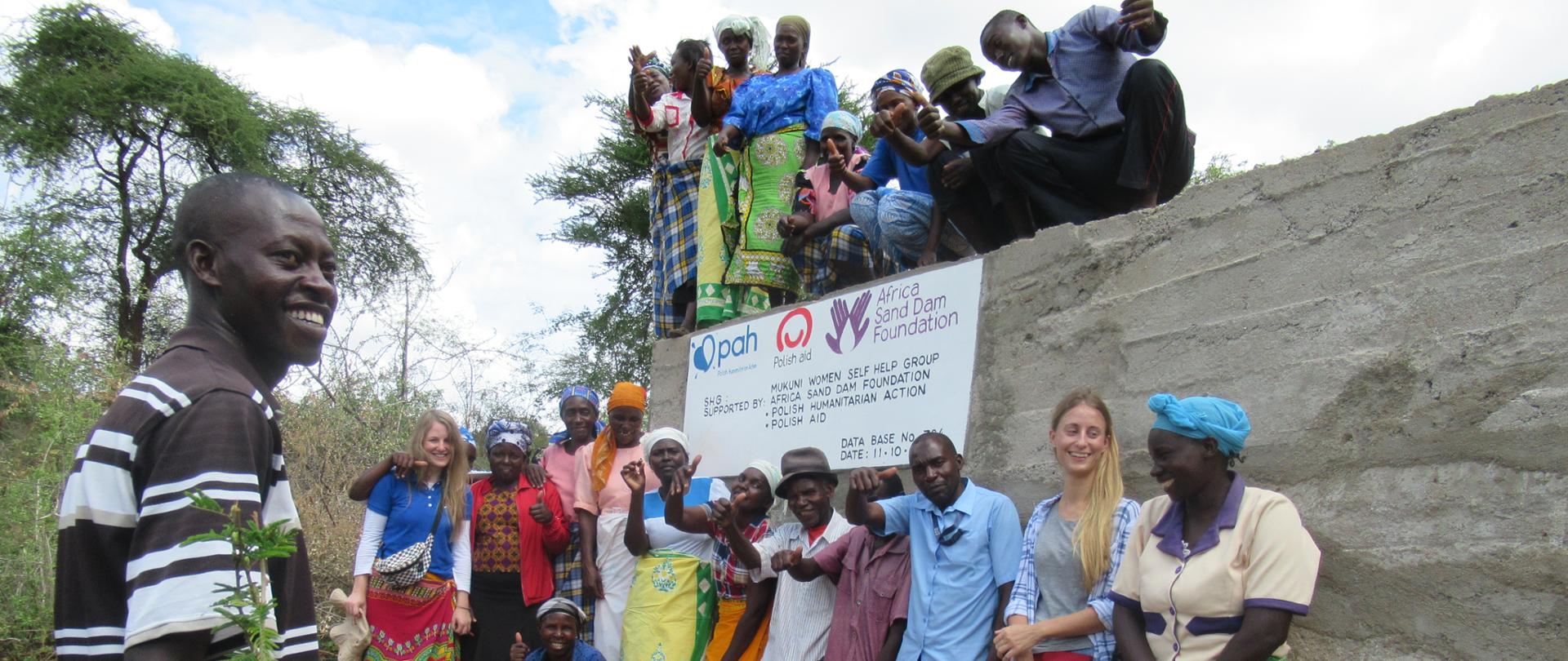 grupa osób stojących przy zbudowanej tamie. Na tamie napis oraz logotypy podmiotów zaanagażowanych w budowę