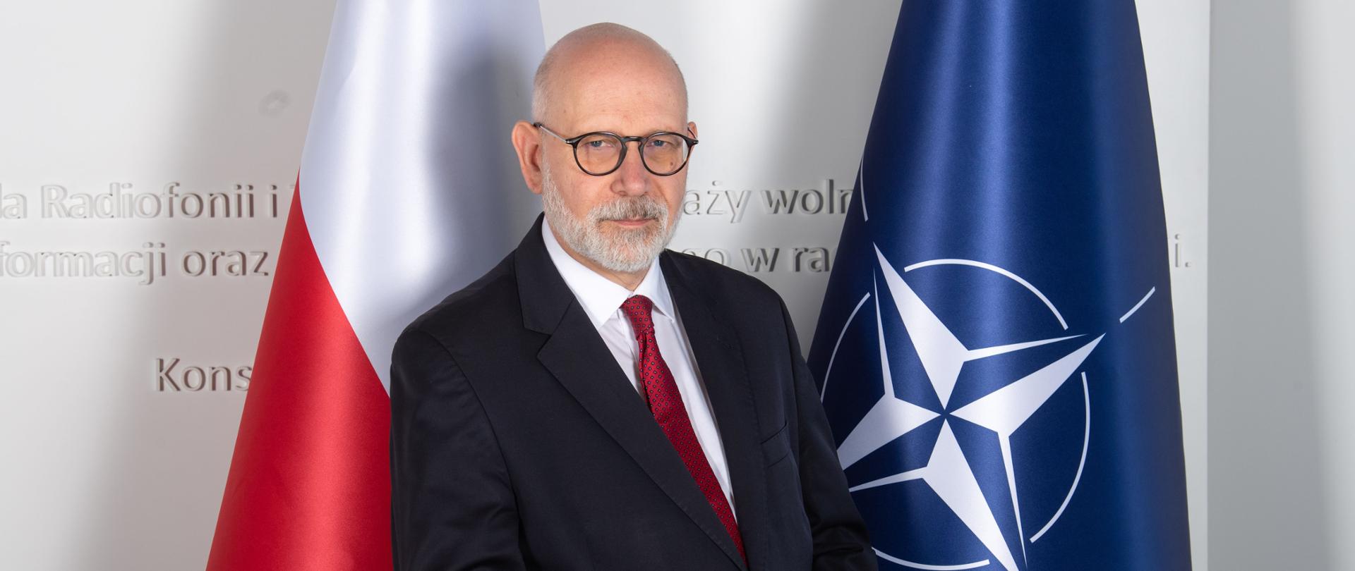 Przewodniczący KRRiT Maciej Świrski. Za nim po lewej stronie flaga biało-czerwona, po prawej stronie niebieska flaga NATO