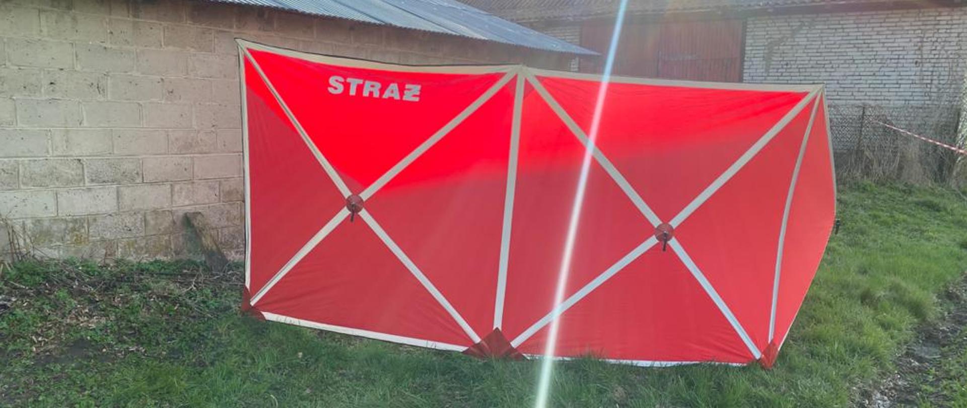 Zdjęcie przedstawia czerwony parawan ochronny osłaniający widok ciała przed osobami postronnymi z białym napisem STRAŻ. Parawan rozstawiony na zielonej trawie przy murowanym budynku gospodarczym.