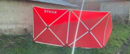Zdjęcie przedstawia czerwony parawan ochronny osłaniający widok ciała przed osobami postronnymi z białym napisem STRAŻ. Parawan rozstawiony na zielonej trawie przy murowanym budynku gospodarczym.