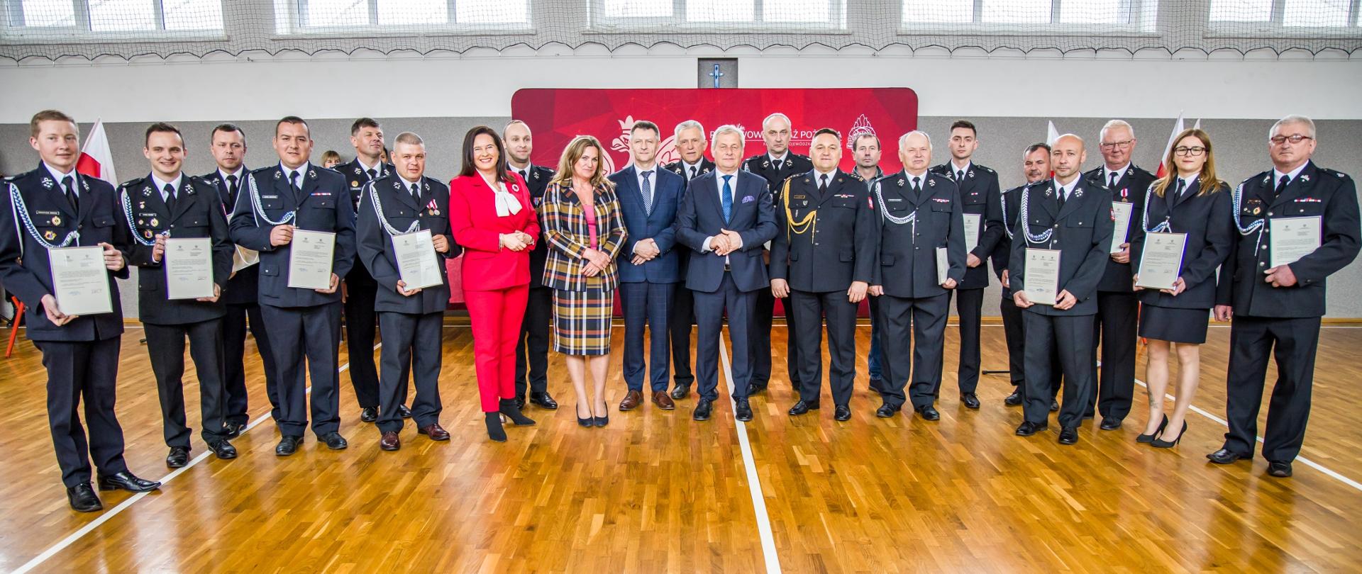 Na zdjęciu uczestnicy uroczystości. Goście oficjalni oraz strażacy OSP z woj. opolskiego. Strażacy trzymają w dłoniach promesy. W tle ścianka z napisem KW PSP. 