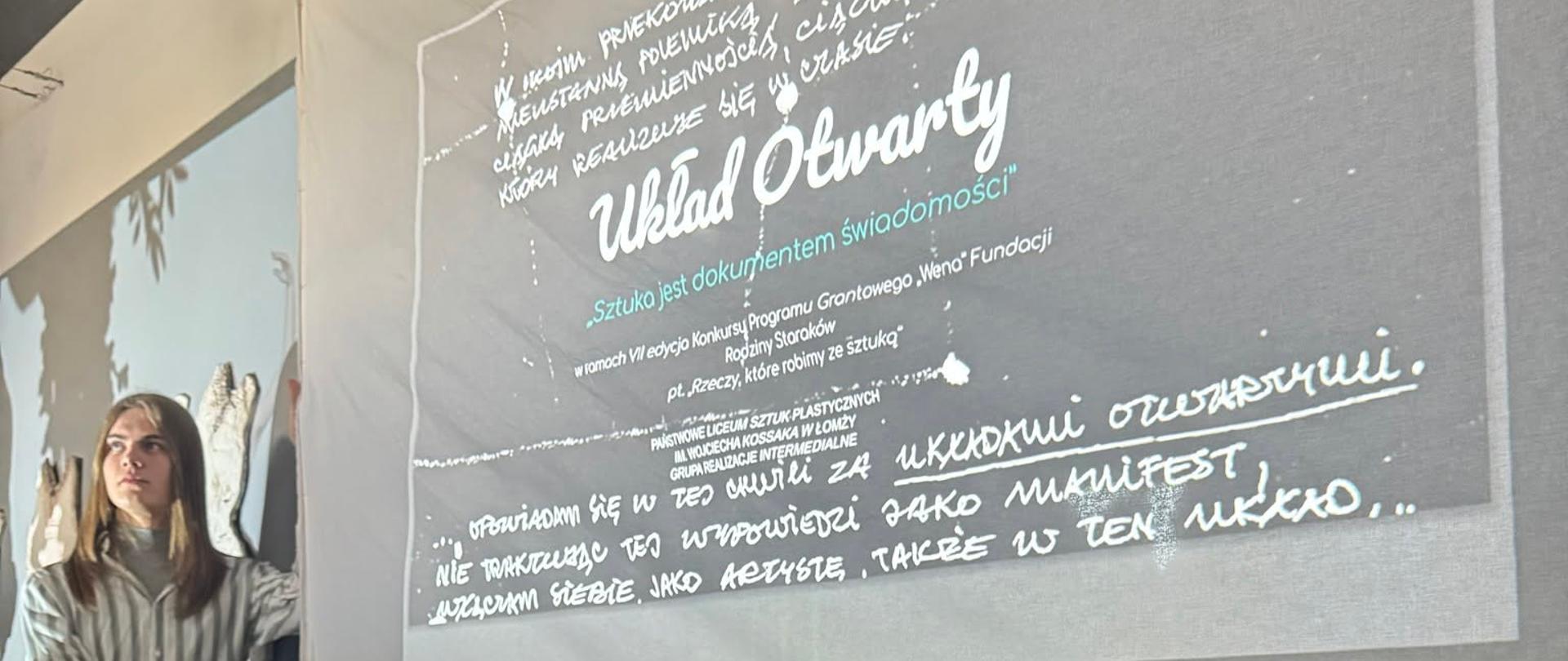 Uczniowie Liceum pod ekranem na którym wyświetlone jest motto wystawy "Układ otwarty - sztuka jest dokumentem tożsamości"
