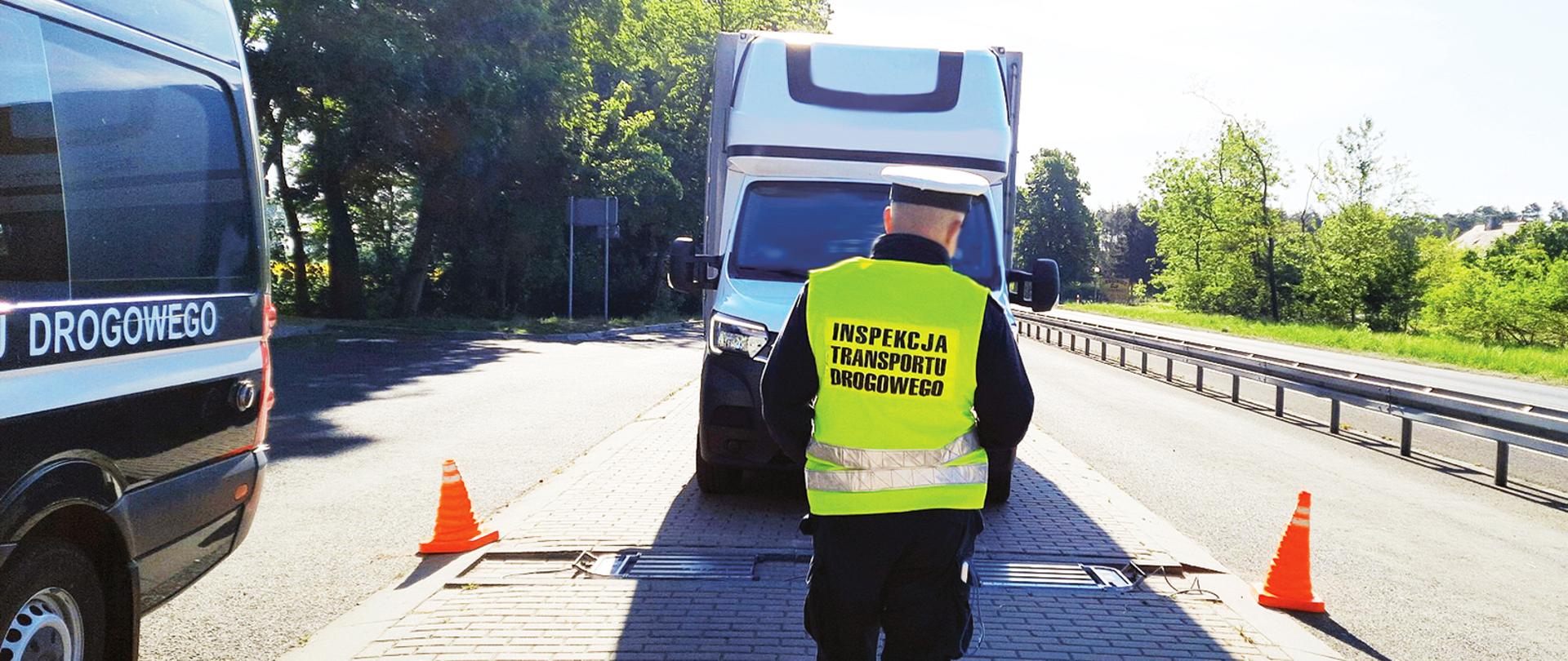 Kontrola konińskich inspektorów wielkopolskiej Inspekcji Transportu Drogowego. W centrum inspektor ITD naprowadza jeden z pojazdów na inspekcyjne wagi. Przed nim, jadący w jego stronę, dostawczak. Po lewej inspekcyjny furgon, po prawej droga.