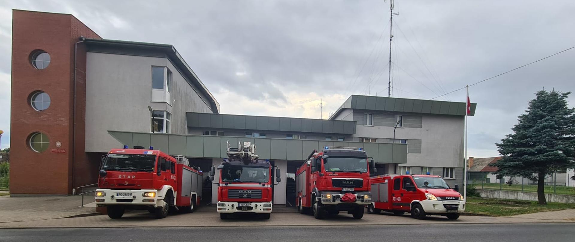 Fotografia przedstawia ujęcie frontu budynku Komendy Powiatowej PSP w Strzelcach Krajeńskich. W czterech otwartych bramach garażu obok siebie wyjeżdżają jednocześnie cztery pojazdy pożarnicze z włączonymi światłami błyskowymi. Jest dzień, niebo jest zachmurzone.