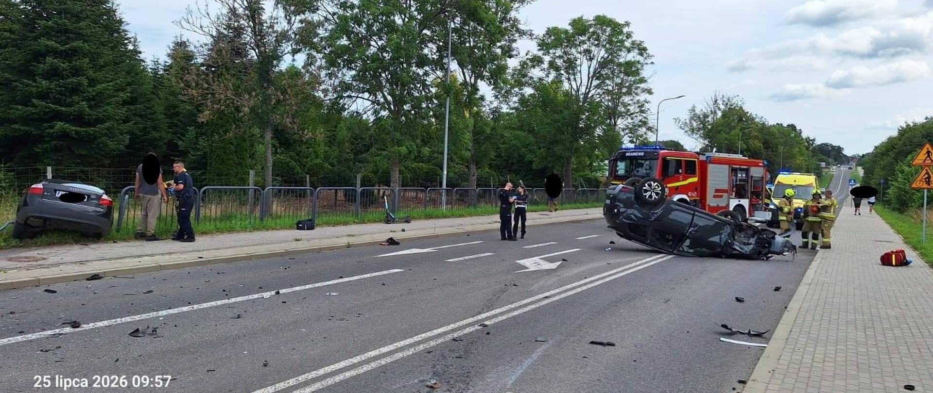 Na zdjęciu widać poważny wypadek drogowy. Jeden samochód leży na dachu, drugi znajduje się przy ogrodzeniu. Na miejscu pracują strażacy, policja i ratownicy medyczni.