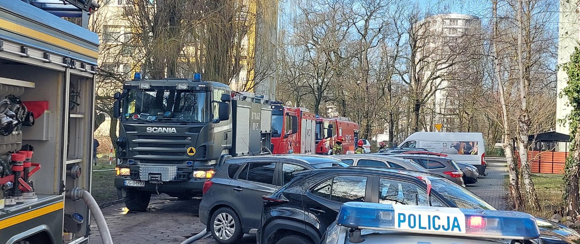 Na zdjęciu widoczne samochody pożarnicze stojące w kolumnie, na froncie samochód koloru zielonego Wojskowej Straży Pożarnej, Następnie dwa czerwone samochody pożarnicze Państwowej Straży Pożarnej. Po prawej stronie widać samochody osobowe zaparkowane prostopadle do chodnika.