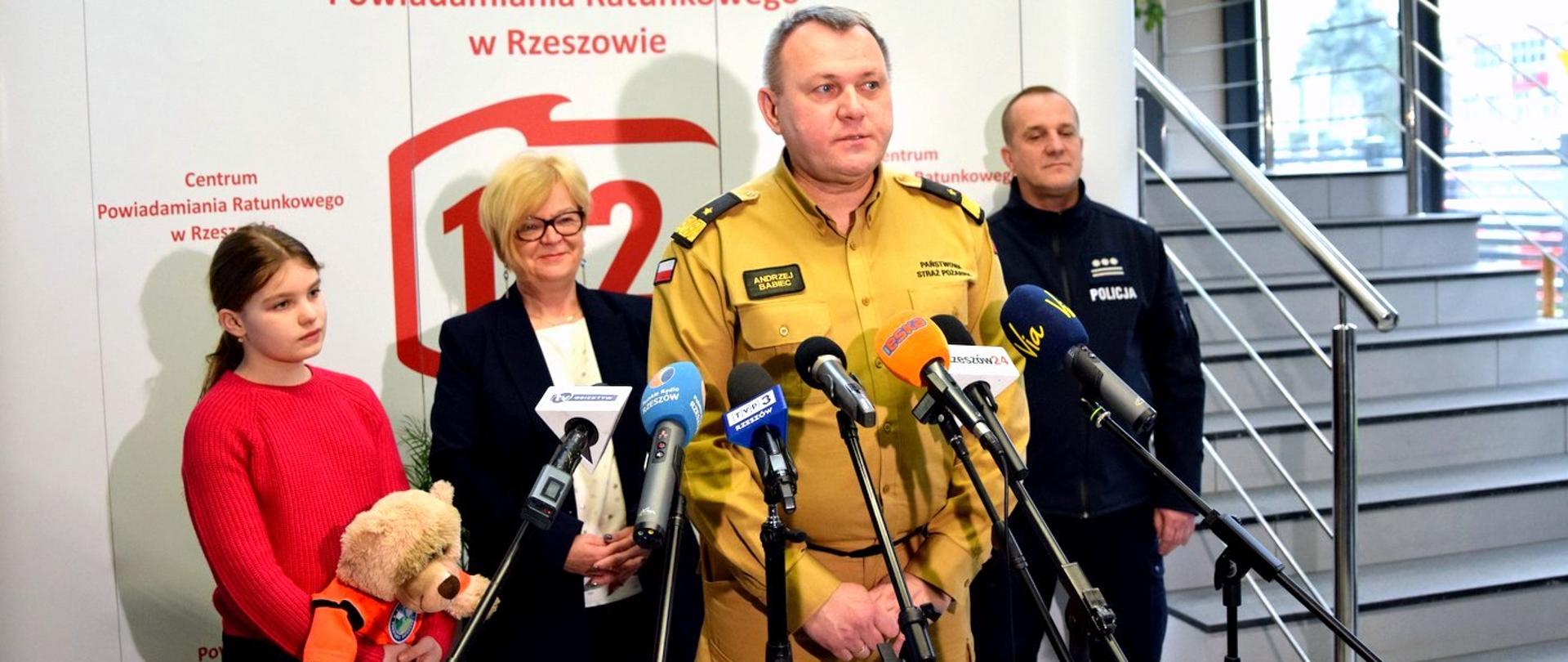 Zdjęcie zrobione w pomieszczeniu, podczas konferencji prasowej z okazji europejskiego dnia numeru alarmowego 112. Przed mikrofonami stoi oficer Państwowej Straży Pożarnej w stopniu generała i przemawia. Za nim ustawieni są pozostali uczestnicy konferencji prasowej.