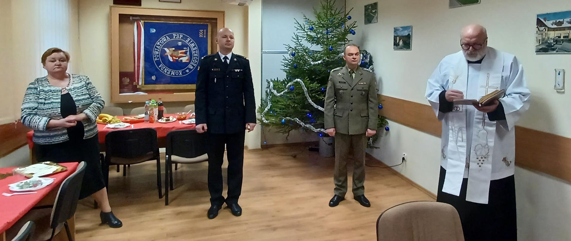 Na świetlicy stoi komendant, dowódca 11 MPA, pani starosta oraz ksiądz który trzyma w rękach pismo święte. Za nimi widać sztandar KP oraz stoi choinka. 