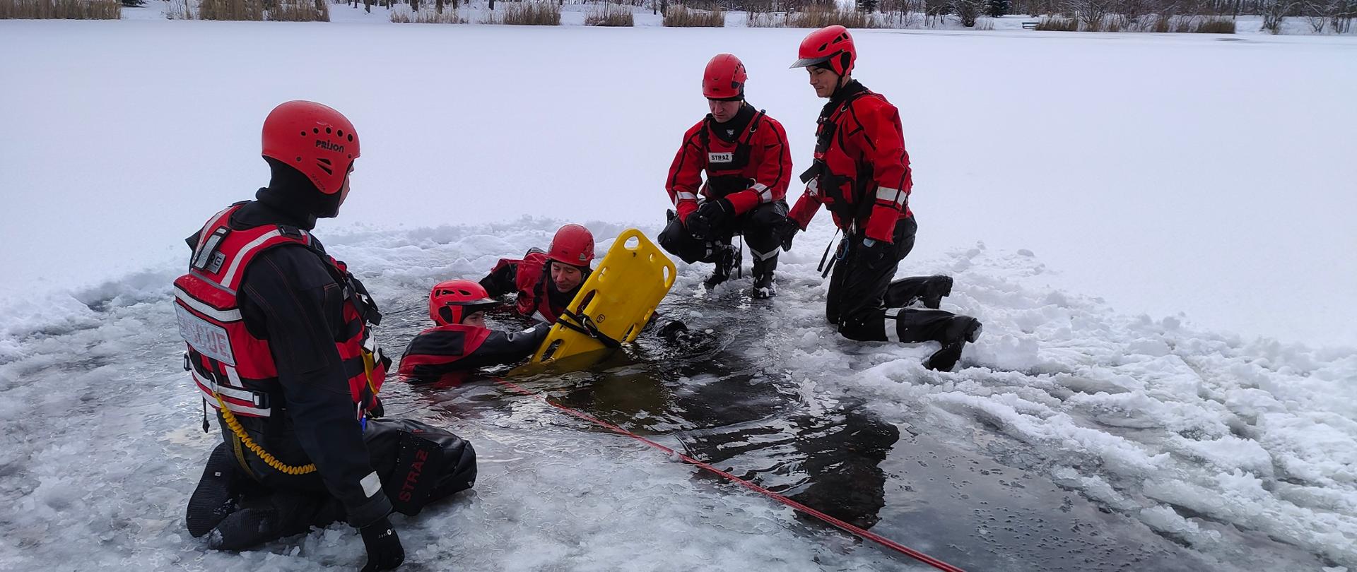 Strażacy na zamarzniętym stawie. Dwóch strażaków znajduje się w wodzie w wyciętym przeręblu.