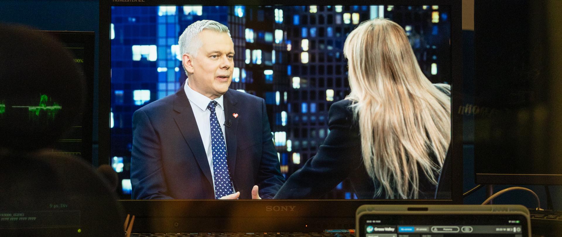 Na zdjęciu widać ekran, który pokazuje widok z kamery w studiu TVN24. Na ekranie dwie osoby - po lewej minister Tomasz Siemoniak, a po prawej redaktor Monika Olejnik (siedząca tyłem). Na dole zdjęcia widać klawiatury, konsole i ekrany.