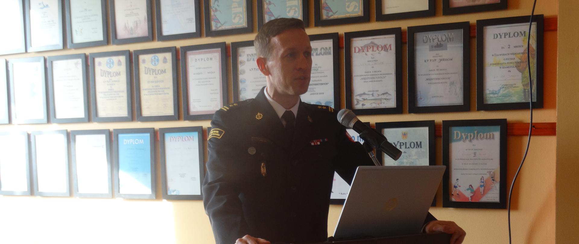 strażak stoi przy mównicy, za nim ściana z dyplomami