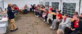 Pod namiotem strażak edukuje grupę dzieci uczestników obozowiska. Strażak prezentuje zagrożenia jakie mogą wystąpić na obozie oraz metody działań. Dzieci zasiadają w ławkach.