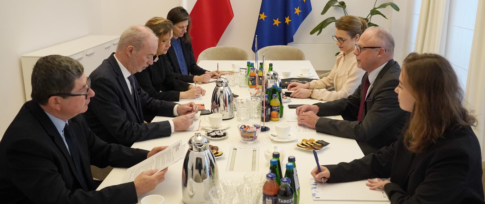 Grupa osób siedzi na wprost siebie przy stole. Przy ścianie flagi Polski i Francji.