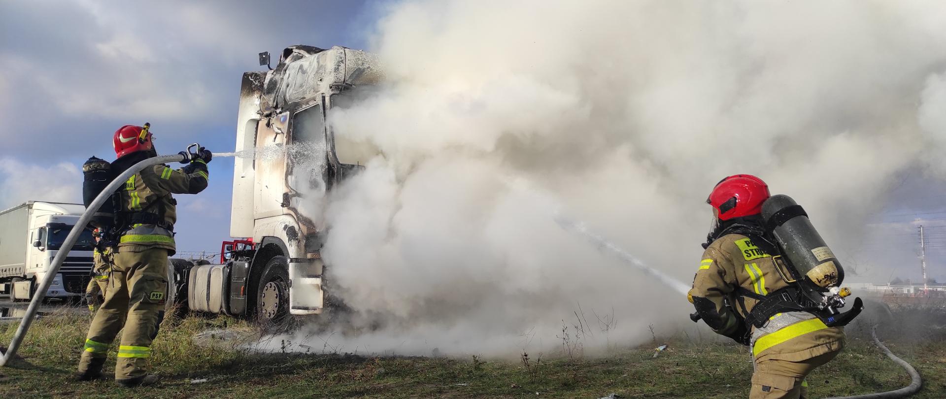 Zdjęcie przedstawia pożar ciągnika siodłowego. Pożarem objęta kabina. Silne zadymienie. Strażacy podają środek gaśniczy na palący się pojazd