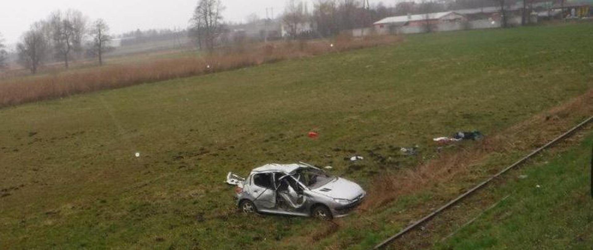 Na zdjęciu widać stojący na łące rozbity srebrny samochód osobowy. Przed pojazdem są tory kolejki wąskotorowej. Obok samochodu na łące leżą rzeczy, które wypadły z samochodu. W tle widać drzewa, krzaki oraz zabudowania. Jest widno.
