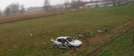 Na zdjęciu widać stojący na łące rozbity srebrny samochód osobowy. Przed pojazdem są tory kolejki wąskotorowej. Obok samochodu na łące leżą rzeczy, które wypadły z samochodu. W tle widać drzewa, krzaki oraz zabudowania. Jest widno.