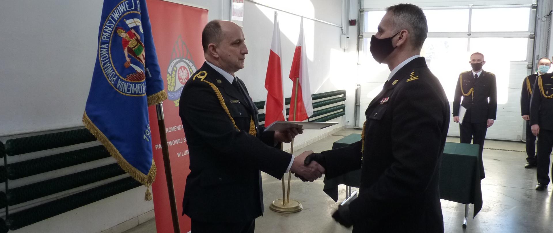 Zdjęcie zrobione w pomieszczeniu strażnicy, na tle Sztandaru Komendy Powiatowej Państwowej Straży Pożarnej w Lublińcu oraz Flagi Państwowej. Na pierwszym planie dwóch strażaków z uściskiem dłoni, zwróconych ku sobie w umundurowaniu galowym.