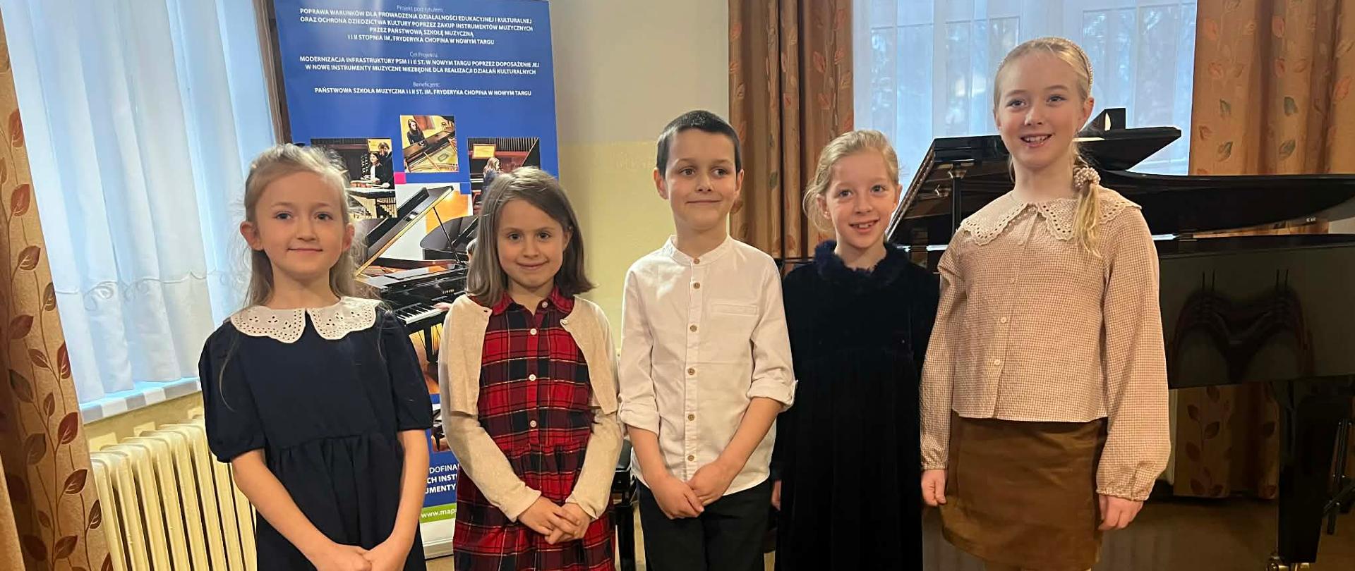 Zdjęcie przedstawia grupę pięciorga uśmiechniętych dzieci w wieku szkolnym, stojących w rzędzie w sali koncertowej lub klasie muzycznej. Postacie: Dzieci są ubrane w eleganckie, wizytowe stroje (sukienki, biała koszula), co sugeruje udział w występie, egzaminie lub uroczystości szkolnej. Tło: Za dziećmi widoczny jest czarny fortepian skrzydłowy oraz baner informacyjny Państwowej Szkoły Muzycznej w Nowym Targu, dotyczący funduszy europejskich na instrumenty muzyczne. Otoczenie: Sala jest jasna, z dużymi oknami przesłoniętymi firankami i klasycznymi zasłonami w liściasty wzór. Na podłodze znajduje się jasna wykładzina.